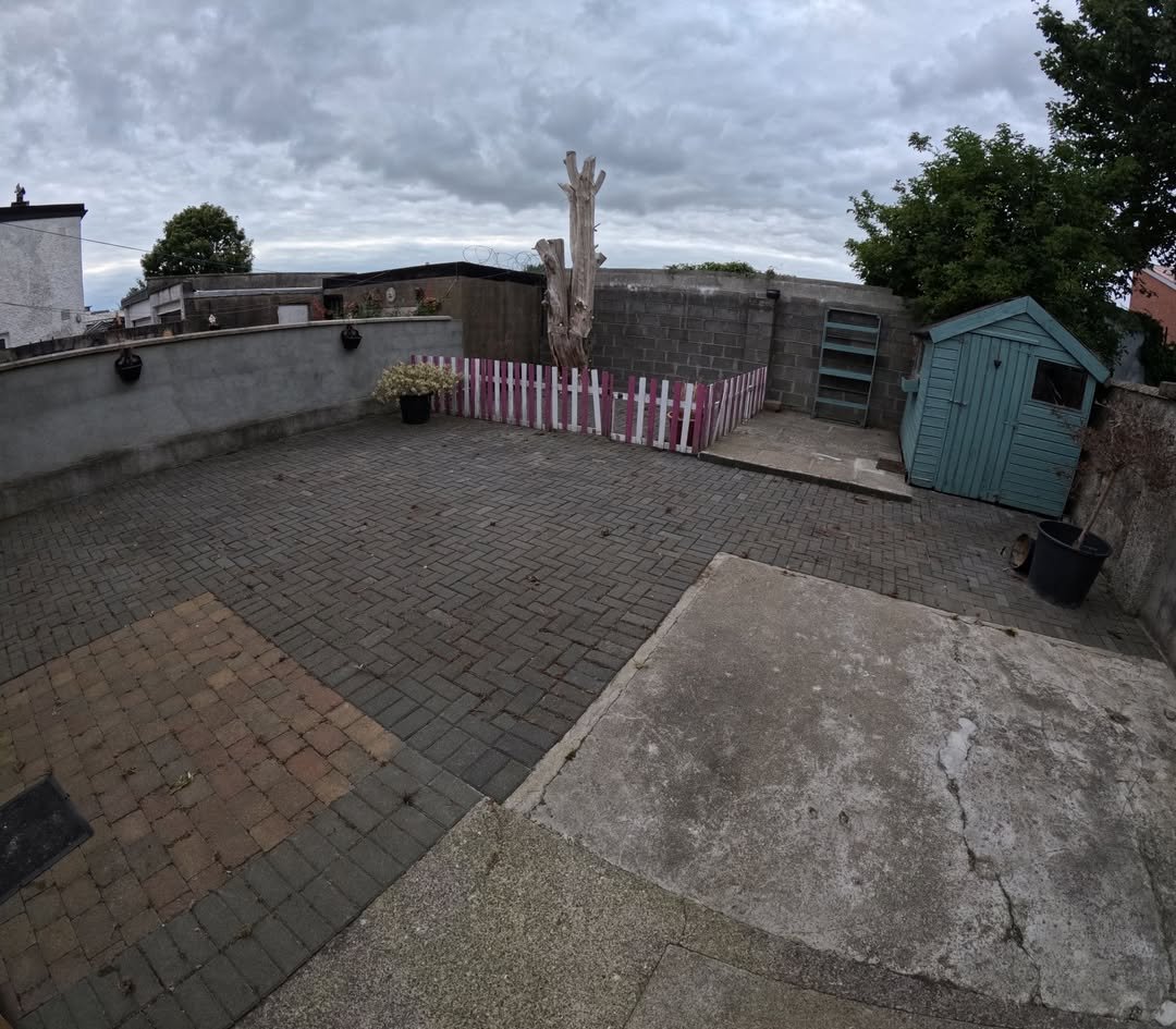 Backyard patio with brick pavers, shed, and pink/white fence under a cloudy sky.