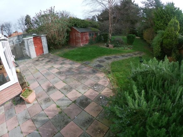 Backyard with patio, shed, grass, and garden. Stone path leads through the green lawn. Overcast day.