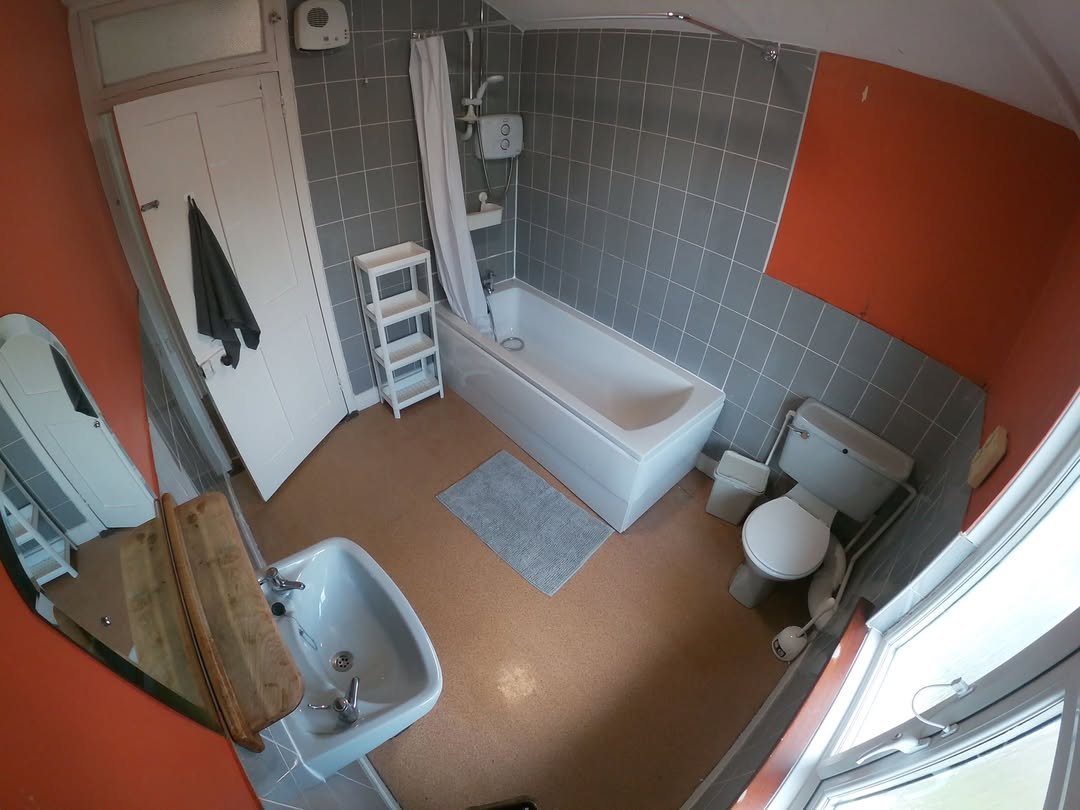 Bathroom with gray tile, white tub, toilet, sink, orange wall, and window. Small gray rug on brown floor.