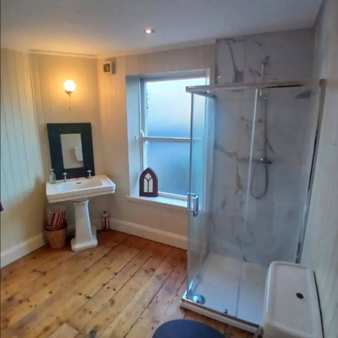 Bathroom with pedestal sink, shower, wood floors, and window.