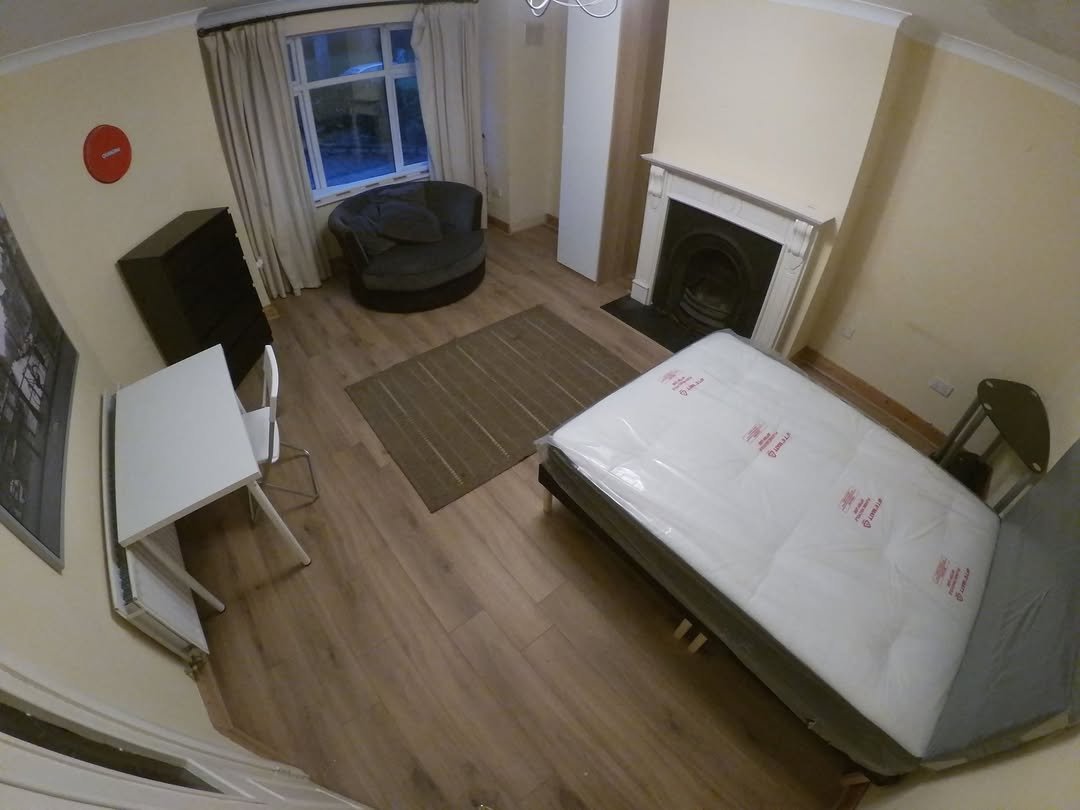 Bedroom with bed, desk, chair, fireplace, and round seat. Neutral colors.