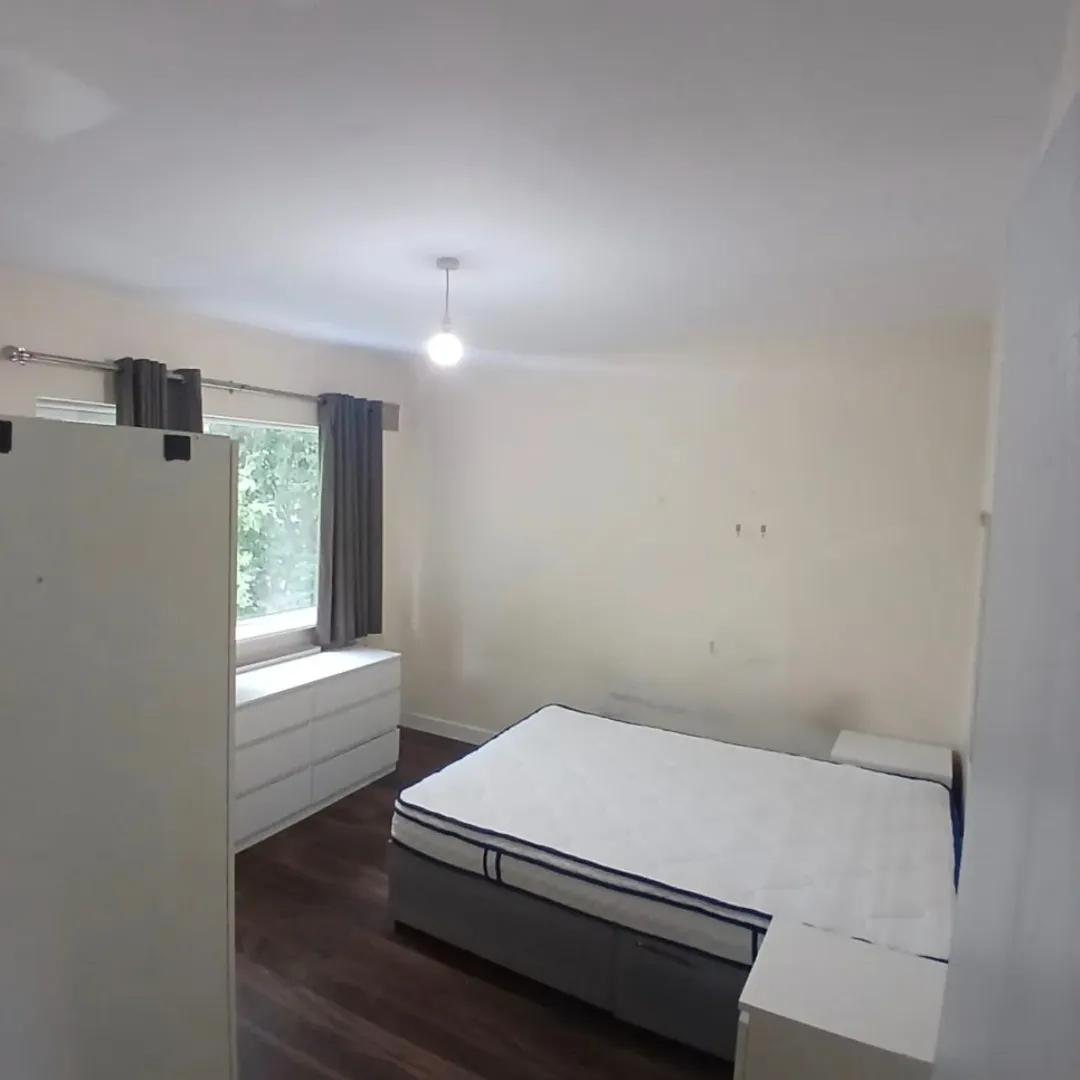 Bedroom with bed, dresser, curtains, and wood floor. Natural light from window.