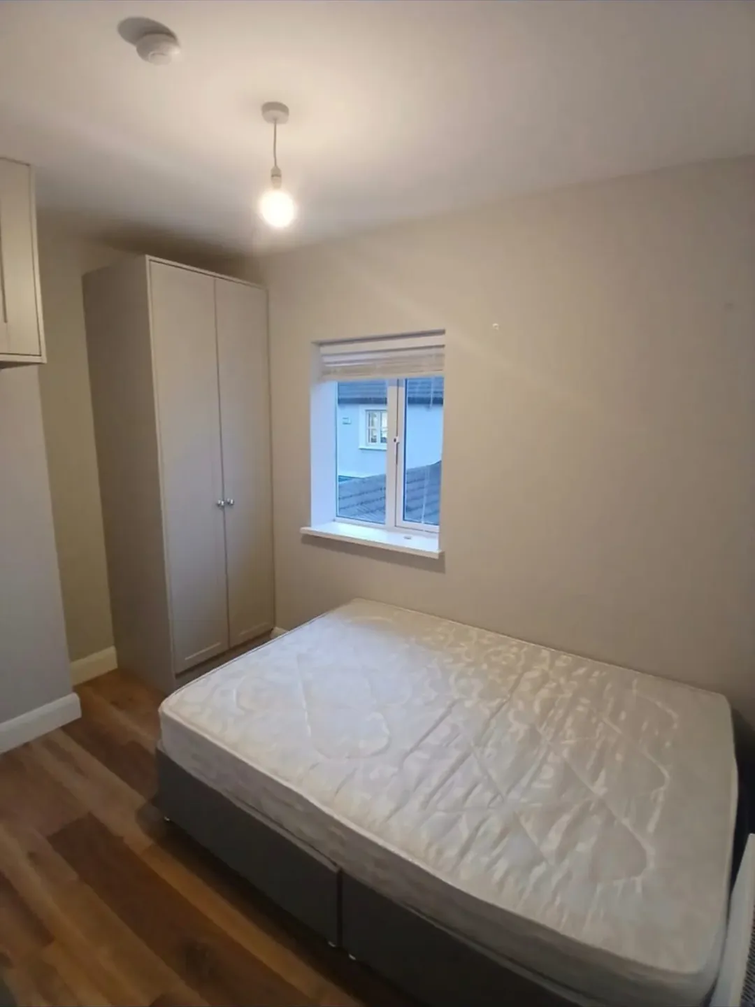 Bedroom with double bed, wardrobe, window, and wood flooring. Neutral color scheme, simple decor.