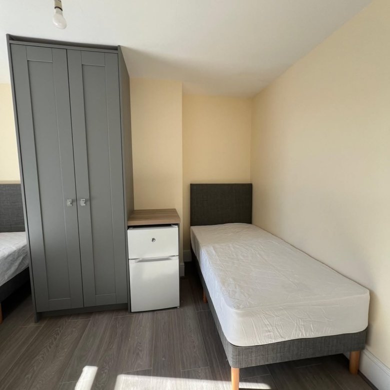 Bedroom with single bed, grey wardrobe, and mini fridge. Neutral tones.
