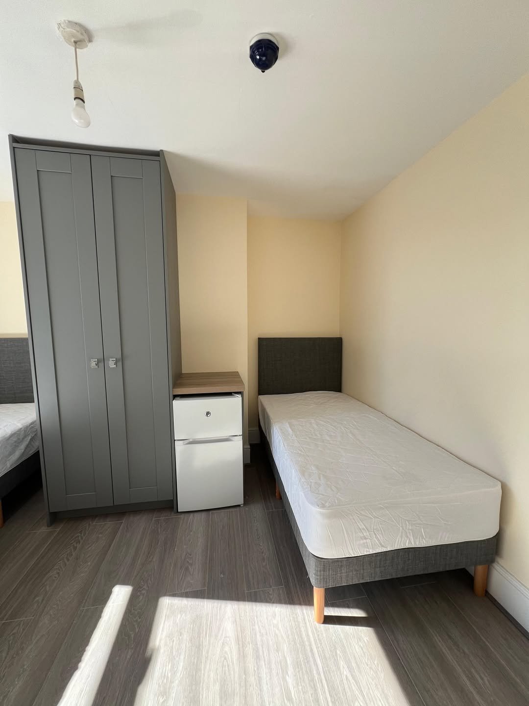 Bedroom with single bed, grey wardrobe, and mini fridge. Neutral tones.