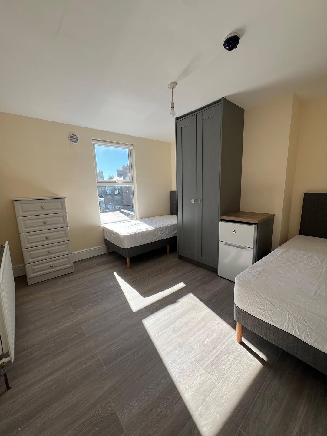 Bedroom with two beds, grey wardrobe and chest of drawers, natural light.