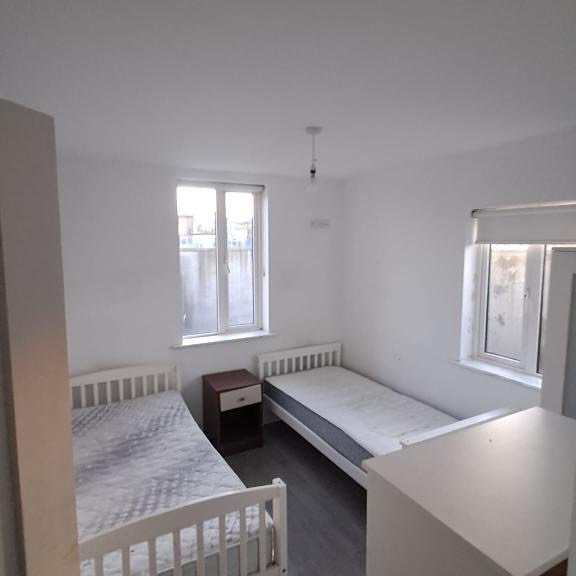 Bedroom with two single beds, nightstand, and natural light from two windows. White walls and gray flooring.