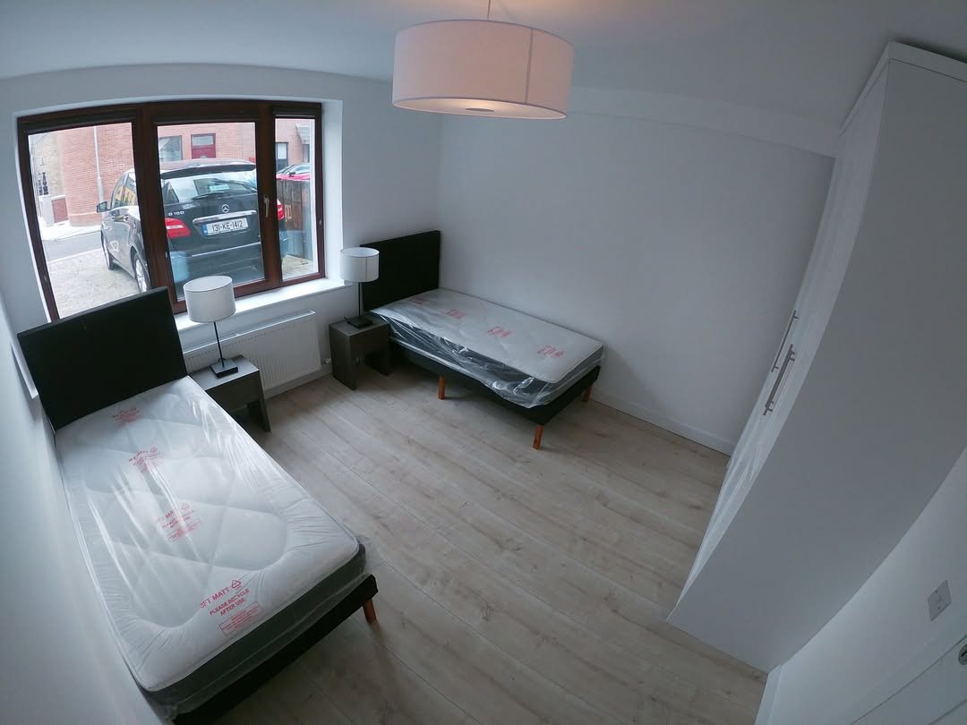 Bedroom with two single beds, bedside tables, wardrobe, and a window view of a car.