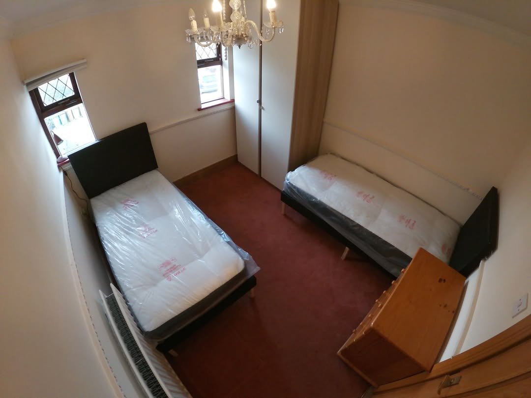 Bedroom with two single beds, wardrobe, chandelier, and window. Red carpet and neutral wall colors.