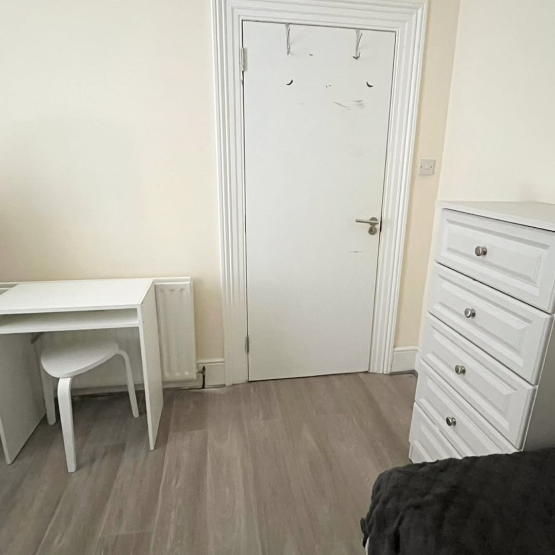 Bedroom corner: white desk, chair, door, and chest of drawers on wood floor.