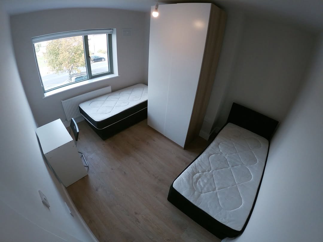 Bright room with two beds, wardrobe, desk, and window. Clean, modern interior.