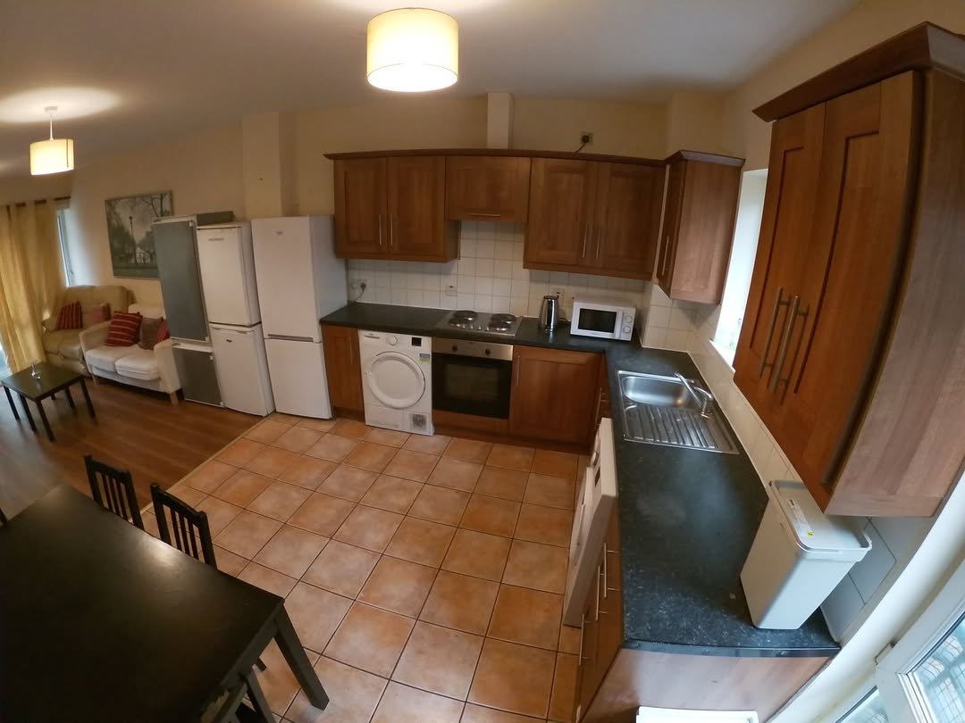 Bright kitchen with appliances, dining table, and living area. Wood cabinets, tile floor.