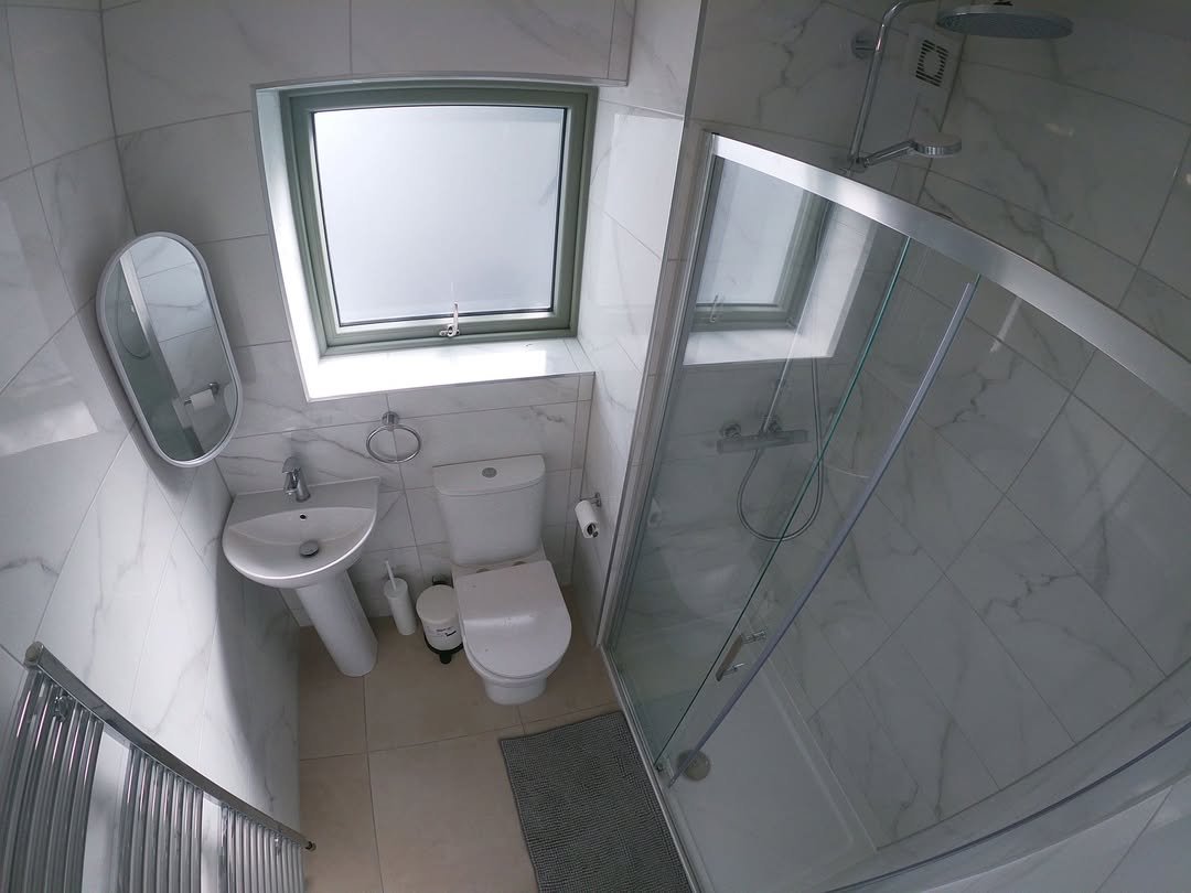 Compact bathroom with shower, toilet, sink, and radiator against white marble-style tiled walls.
