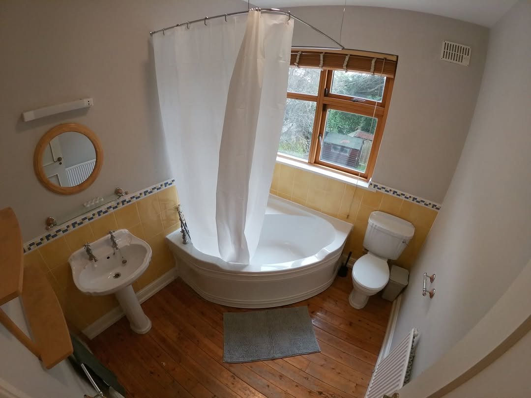 Corner bathtub with shower curtain, pedestal sink, toilet, and wood floor in bathroom.