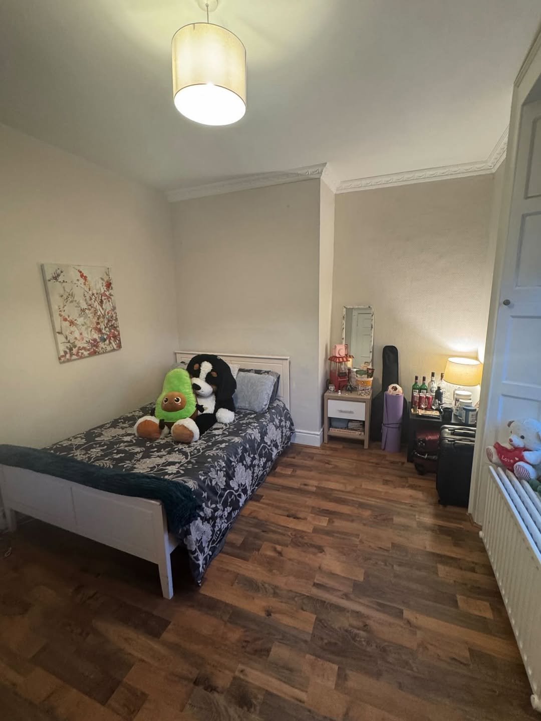 Cozy bedroom with bed, stuffed animals, bedside table, and personal items. Warm lighting and wooden floor.