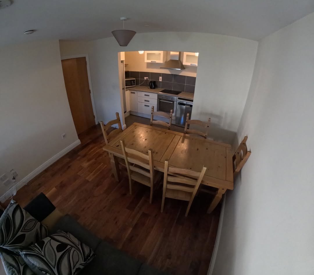Dining area with wooden table and chairs, open kitchen in background.