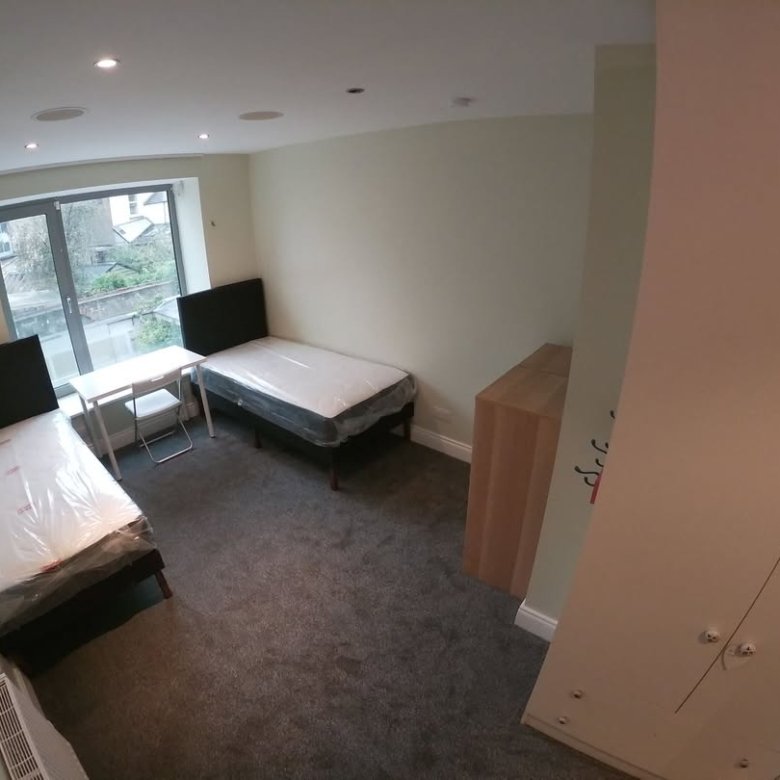 Cozy dorm room with two beds, desk, wardrobe, and carpet. Natural light from the window.