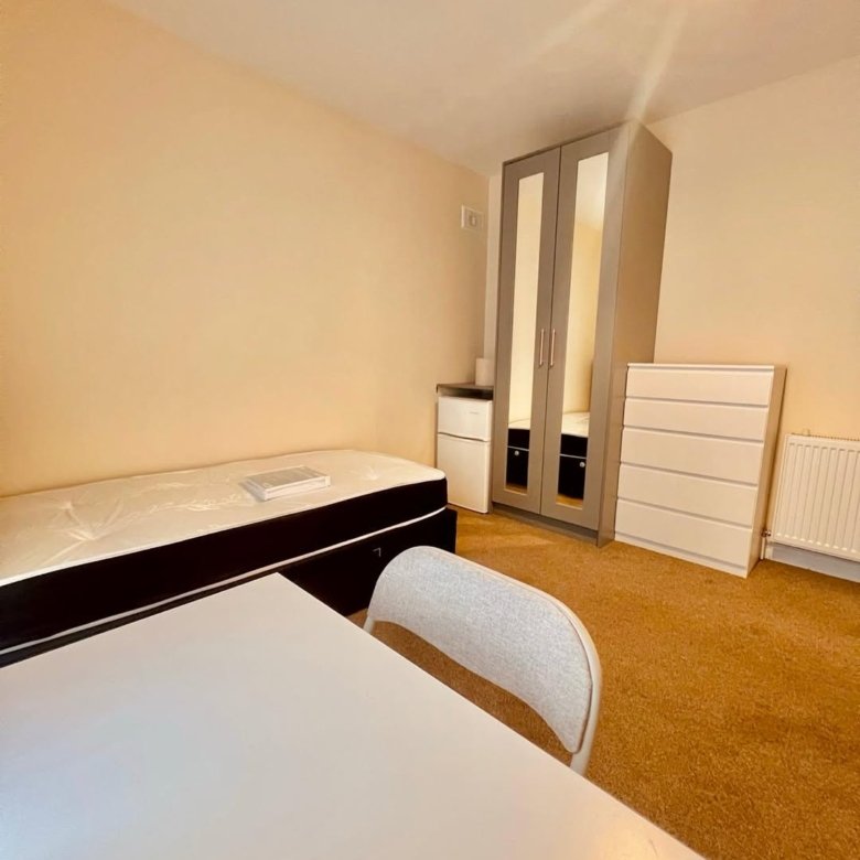 Bedroom with bed, wardrobe, dresser, table, and chair. Neutral color scheme.