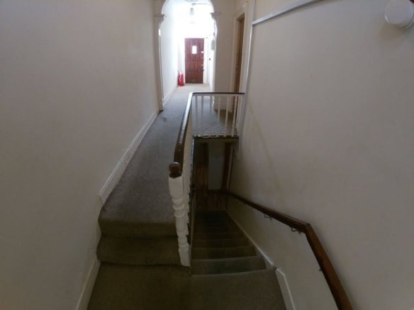Hallway with stairs leading down, carpeted floor, neutral walls, residential building.