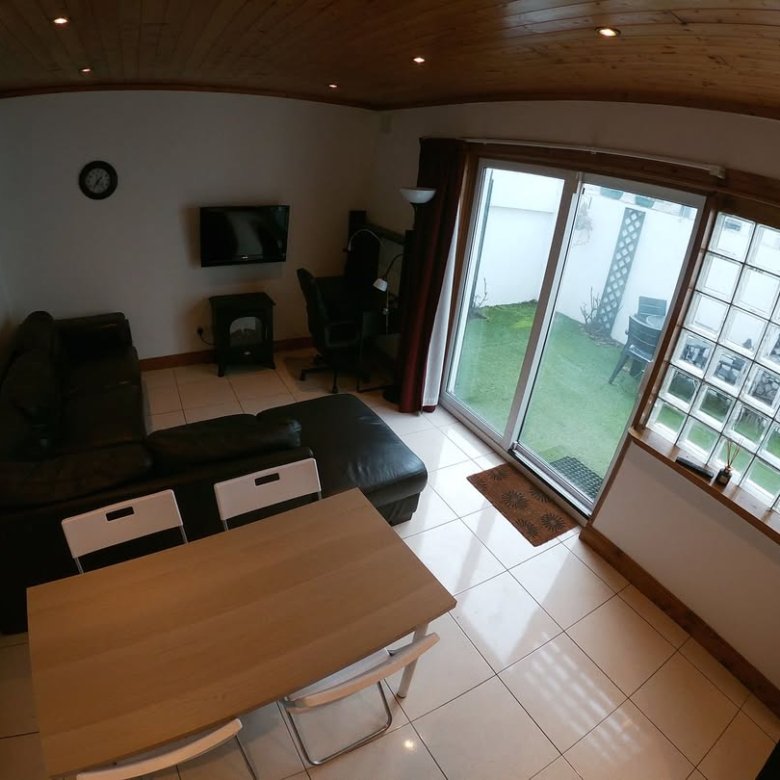 Living room with black leather sofa, dining table, TV, and sliding glass doors to a garden. Natural light and wood accents.