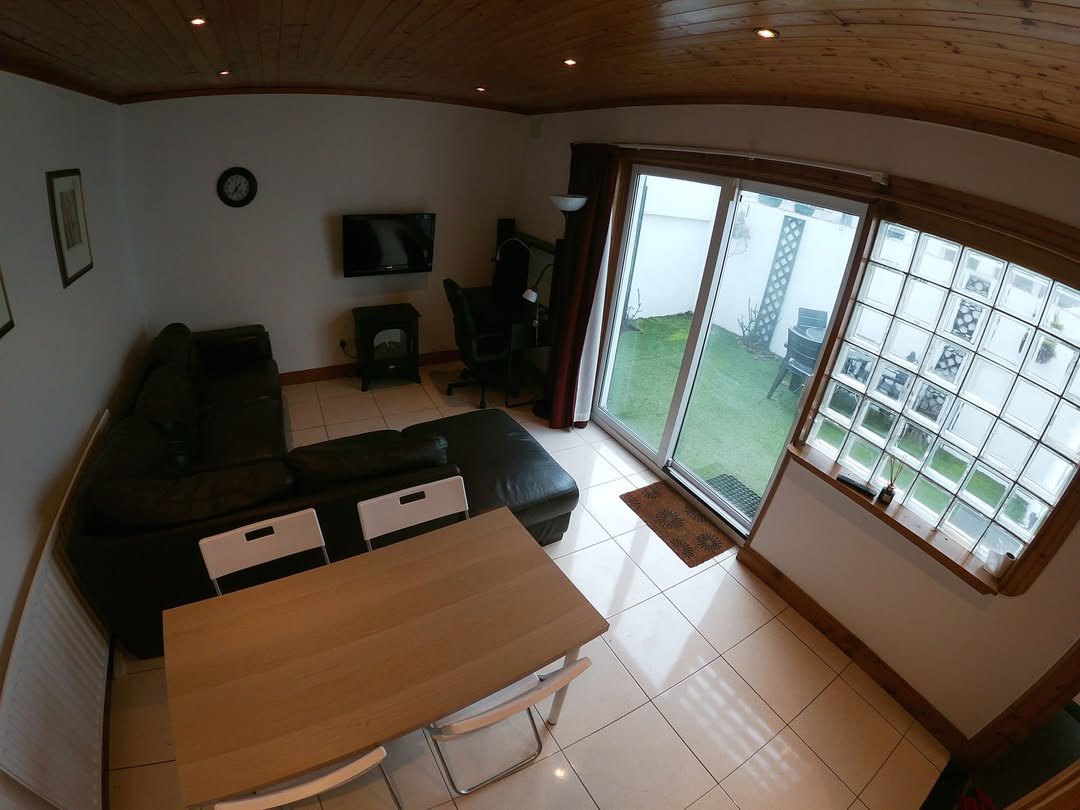 Living room with black leather sofa, dining table, TV, and sliding glass doors to a garden. Natural light and wood accents.