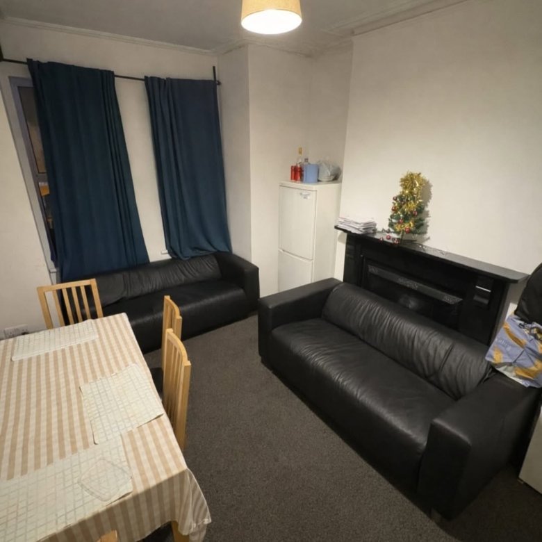 Living room with black sofas, dining table, mini fridge, and small Christmas tree on a fireplace mantel.