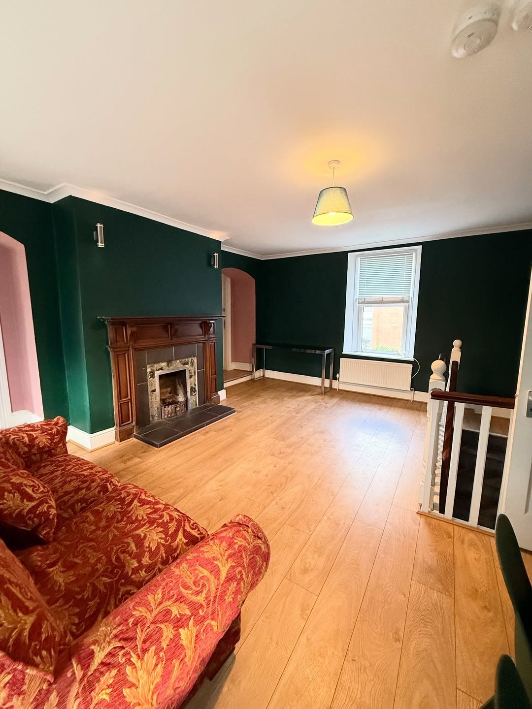 Living room with dark green walls, fireplace, wood floors, and red floral sofa. Bright and airy space.