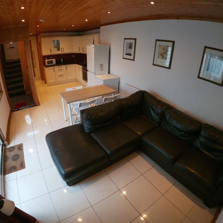 Living room with dark green L-shaped sofa, dining table, kitchen, and stairs. White walls and tile flooring.