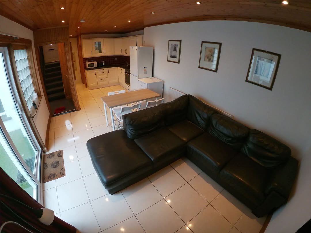 Living room with dark green L-shaped sofa, dining table, kitchen, and stairs. White walls and tile flooring.