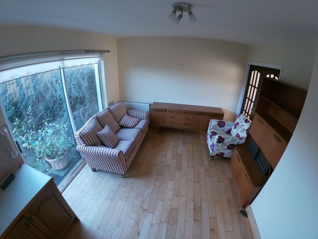 Living room interior with striped sofa, floral armchair, wooden cabinet, and large window overlooking greenery.