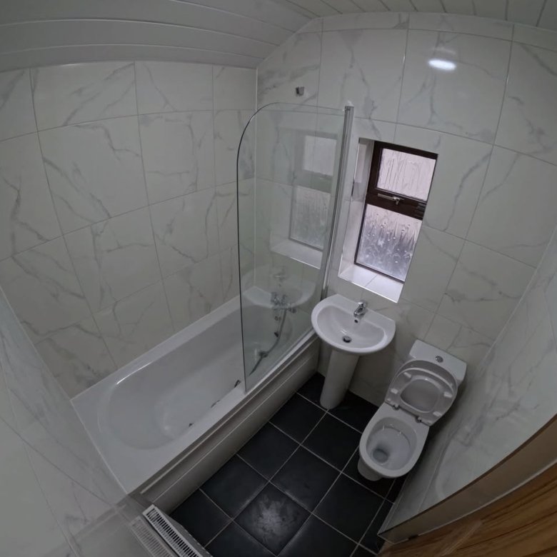 Modern bathroom with tub, shower screen, pedestal sink, and toilet. White tile walls, dark floor.
