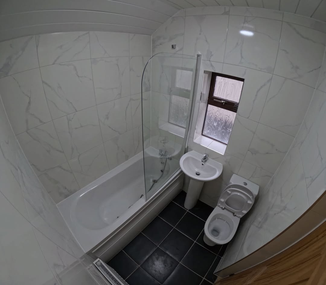 Modern bathroom with tub, shower screen, pedestal sink, and toilet. White tile walls, dark floor.