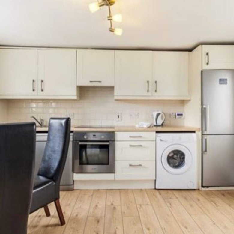 Modern kitchen with cream cabinets, stainless steel appliances, and light wood flooring. Dining chairs visible.