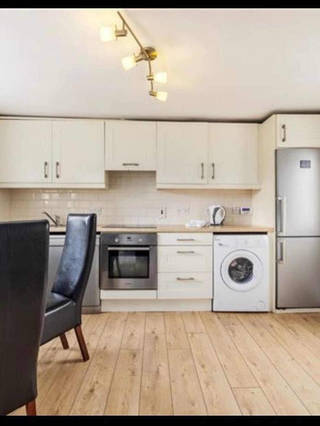 Modern kitchen with cream cabinets, stainless steel appliances, and light wood flooring. Dining chairs visible.