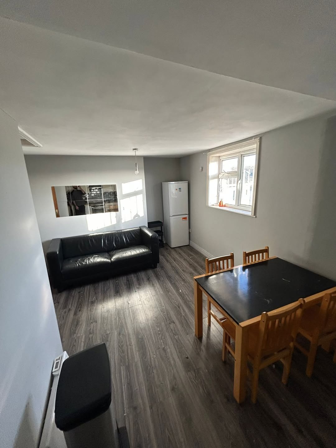 Living room with black sofa, dining table, fridge, and window with natural light. Modern interior design.