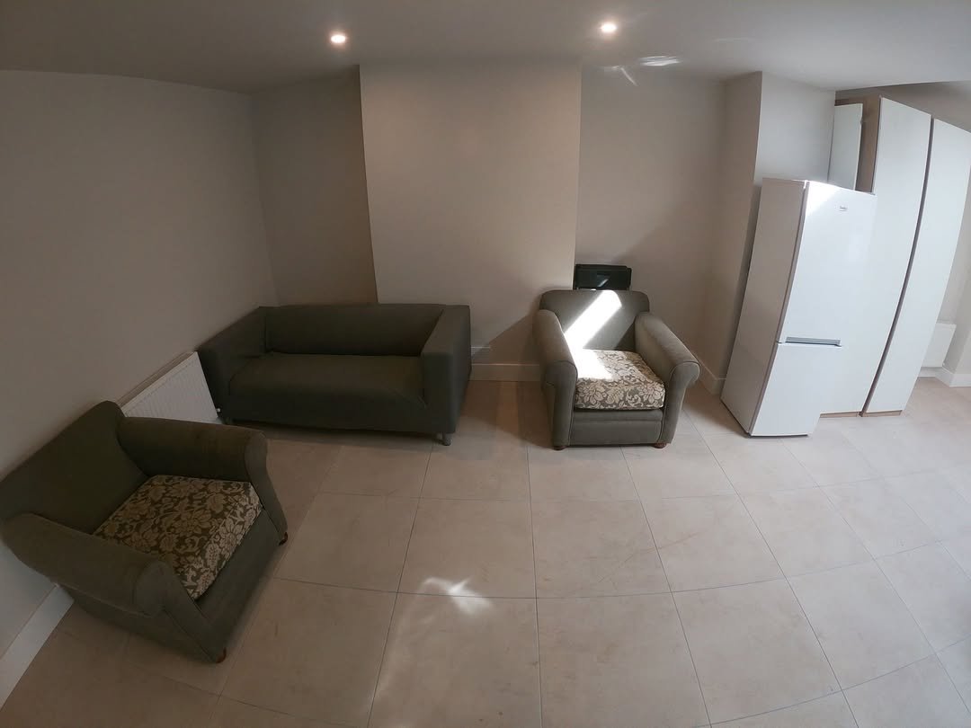 Living room with olive green sofa and armchair, floral cushions, and white refrigerator in a bright, tiled space.