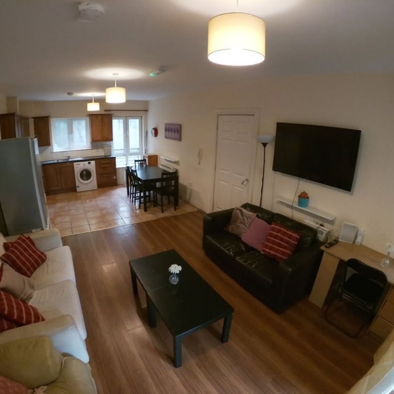 Open-plan apartment interior with living room, dining area, and kitchen. Sofa, table, TV visible.