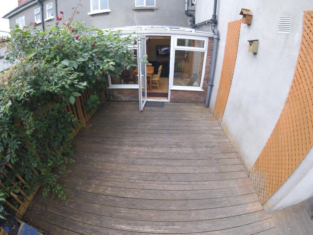 Outdoor patio with wooden deck, greenery, and open door to sunroom.