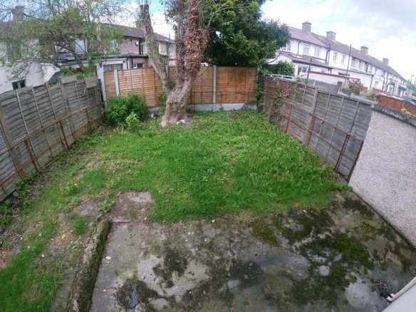 Overgrown backyard with grass, weeds, concrete patio, and wooden fences surrounding the space.