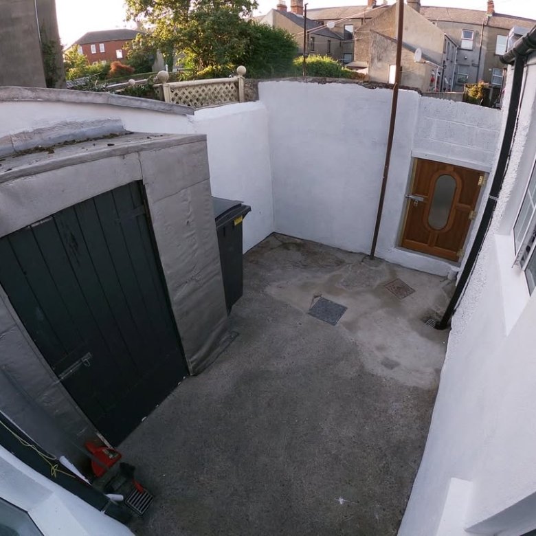 Small backyard with a shed, door to house, and bin, painted white walls, concrete floor, and houses in background.