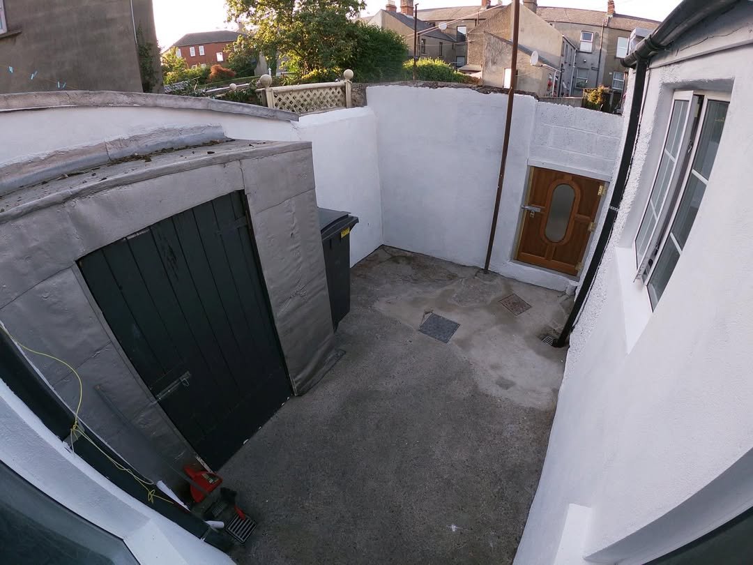 Small backyard with a shed, door to house, and bin, painted white walls, concrete floor, and houses in background.