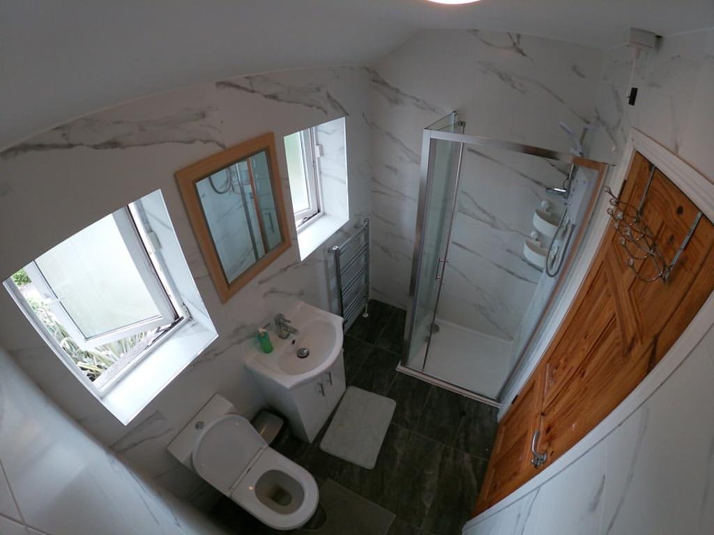 Small bathroom with marble walls, shower, toilet, sink, wood door, and two windows.