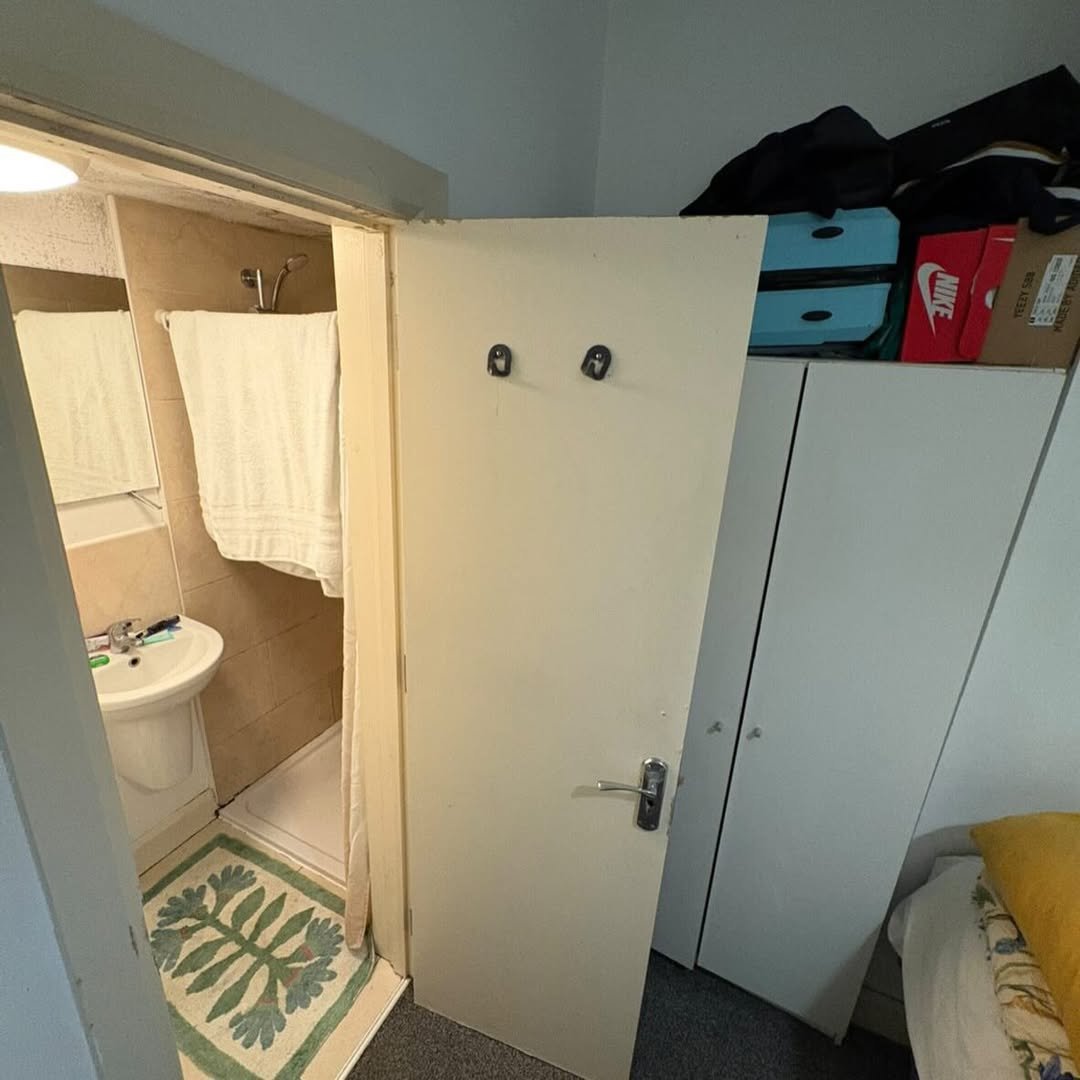 Small bathroom with shower, sink, and open door next to wardrobe and bed.