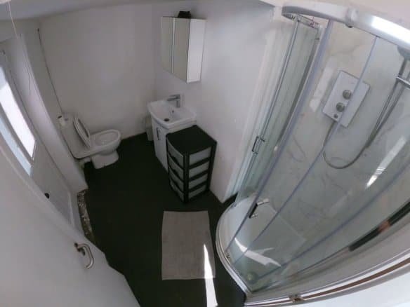 Small bathroom with shower, toilet, sink, and storage unit. White walls and dark floor.
