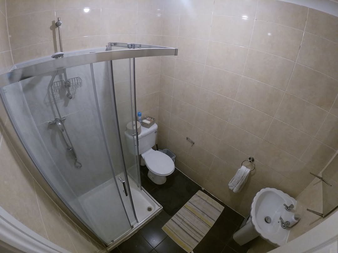 Small bathroom with shower stall, toilet, and sink. Beige tile walls and dark floor.
