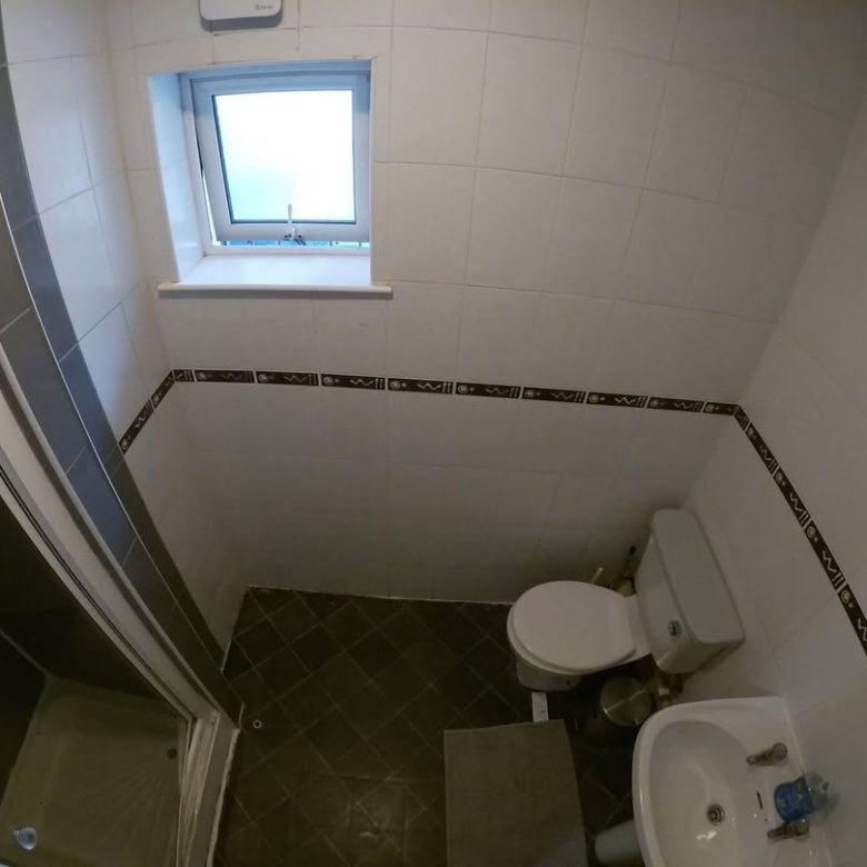 Small bathroom with shower, toilet, and sink beneath a window. Dark tile flooring.