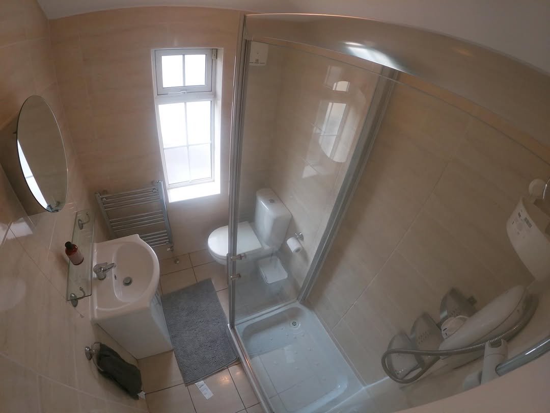 Small bathroom with shower, toilet, sink, and window. Neutral tile walls and gray rug.