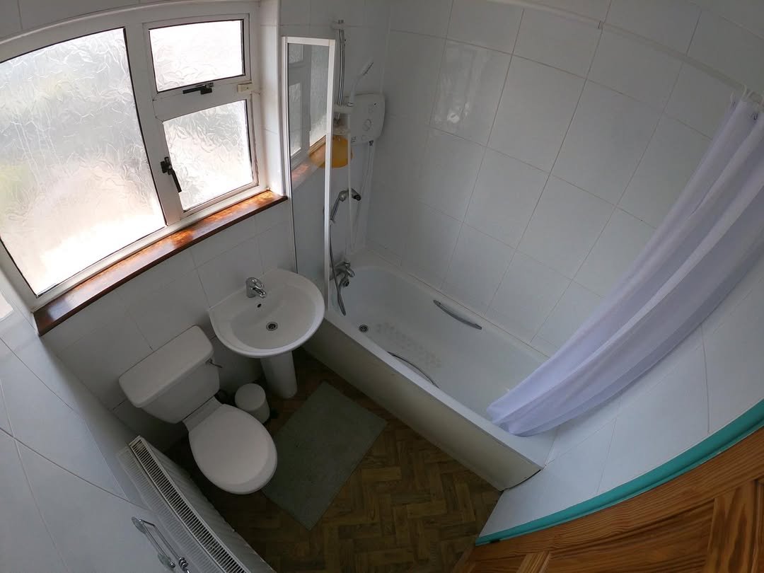 Small bathroom with toilet, sink, bathtub and shower. Window provides natural light. White tile and simple fixtures.