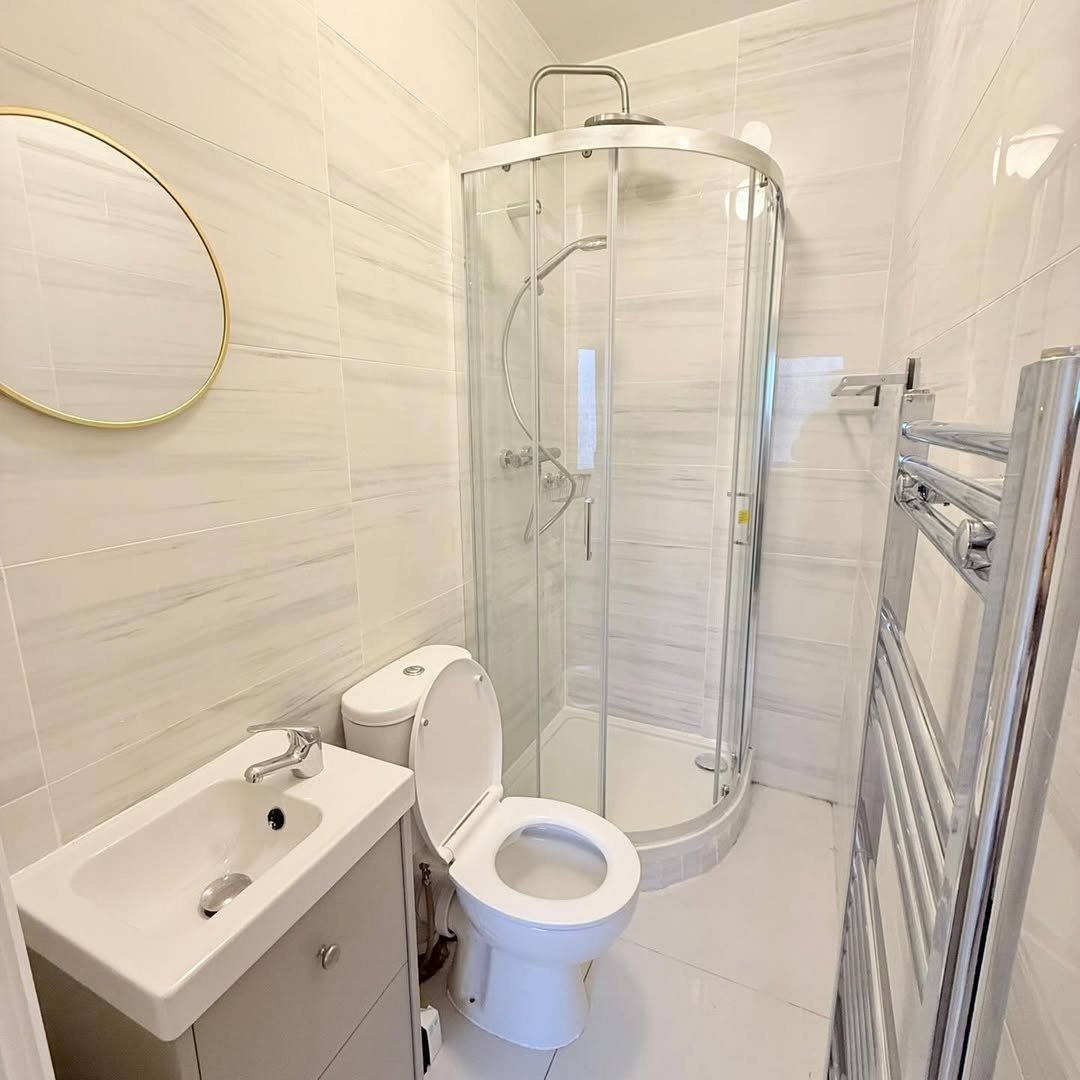 Small bathroom with toilet, sink, shower, and towel warmer. Neutral tile and modern fixtures.
