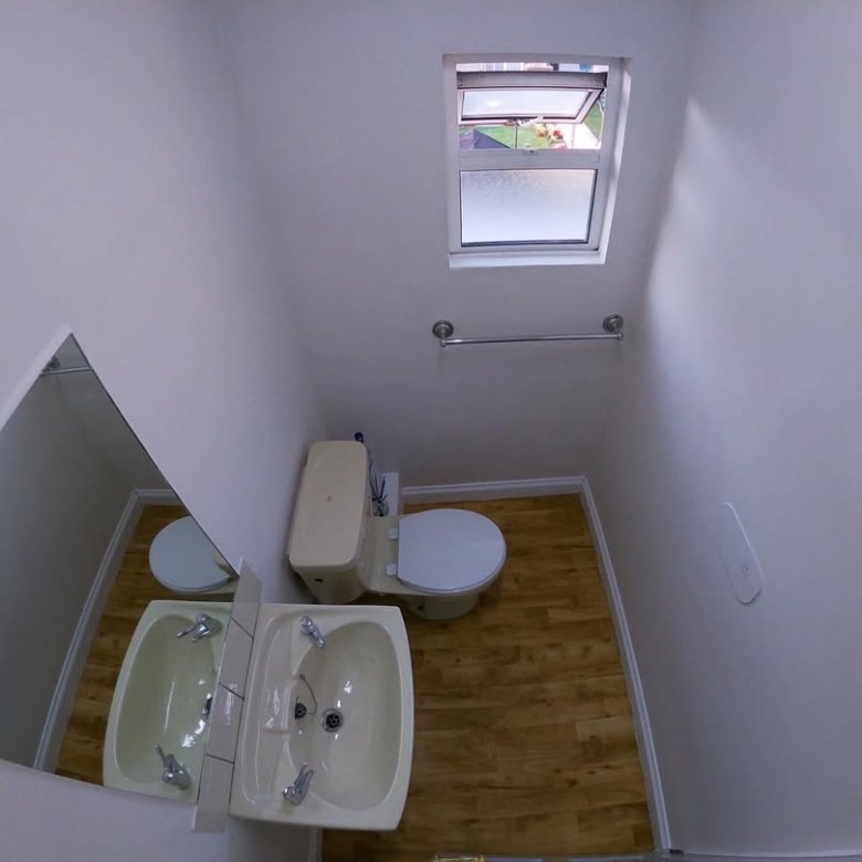 Small bathroom with toilet, sink, mirror, and window, viewed from above. White walls, wood floor.